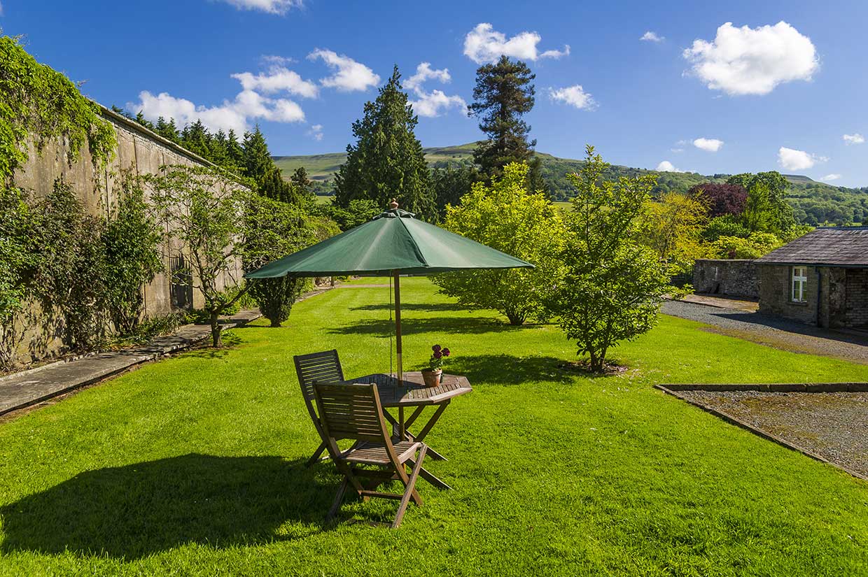 Garden table and chairs beneath a green parasol on a lawn beside a stone wall and cottage at Glanusk Estate