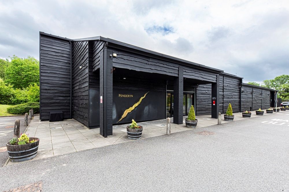 an exterior shot of teh ditillery in Penderryn, dark wooden clad building with a gold splash of their logo non the wall.