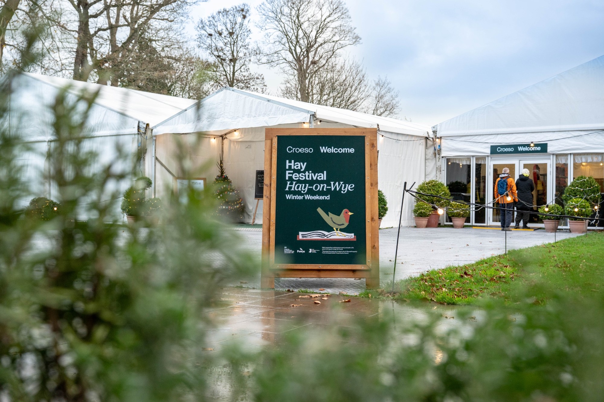 Hay Festival Winter Weekend welcome sign beside marquees, Christmas trees and visitors