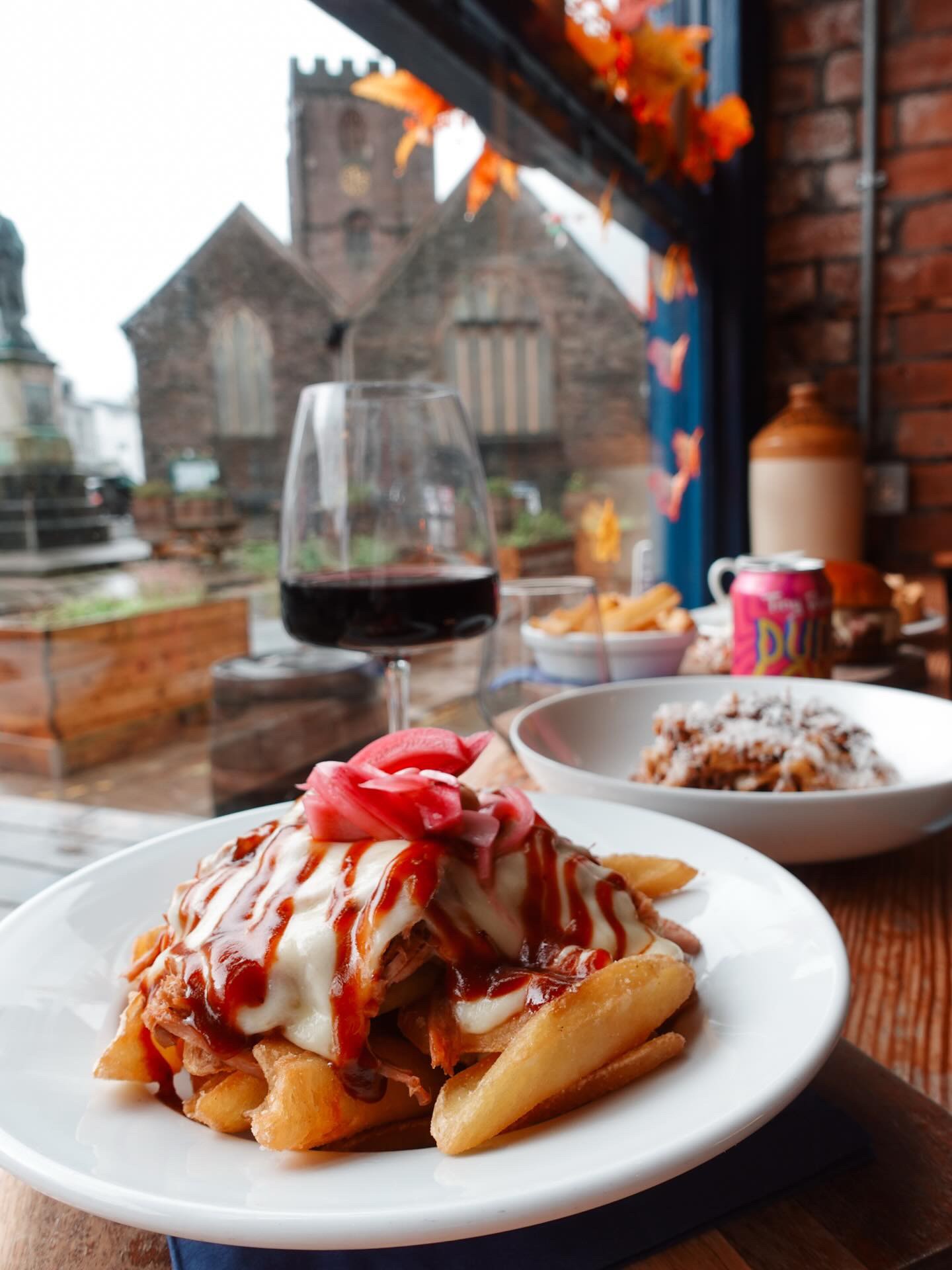 “Loaded fries topped with melted cheese and pickled onions, served by a window at Brecon Tap with Brecon church visible outside