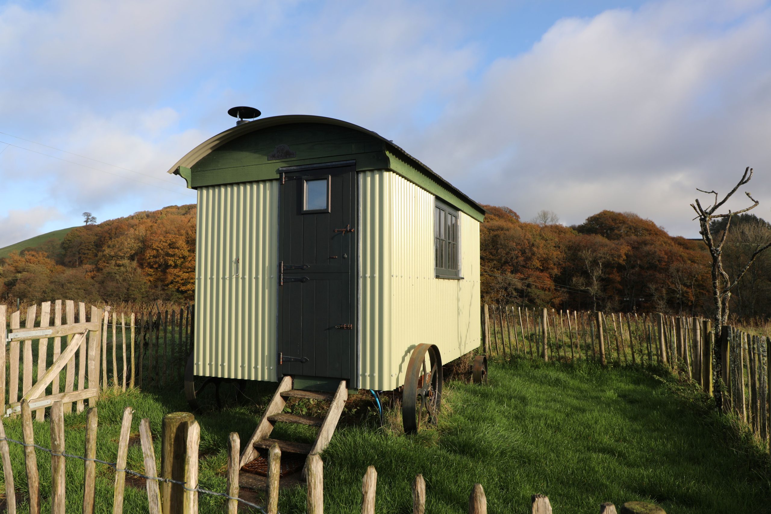 Green shepherd’s hut at Farmstead and Wild, set in a grassy field with autumn woodland and hills in Bannau Brycheiniog (Brecon Beacons).
