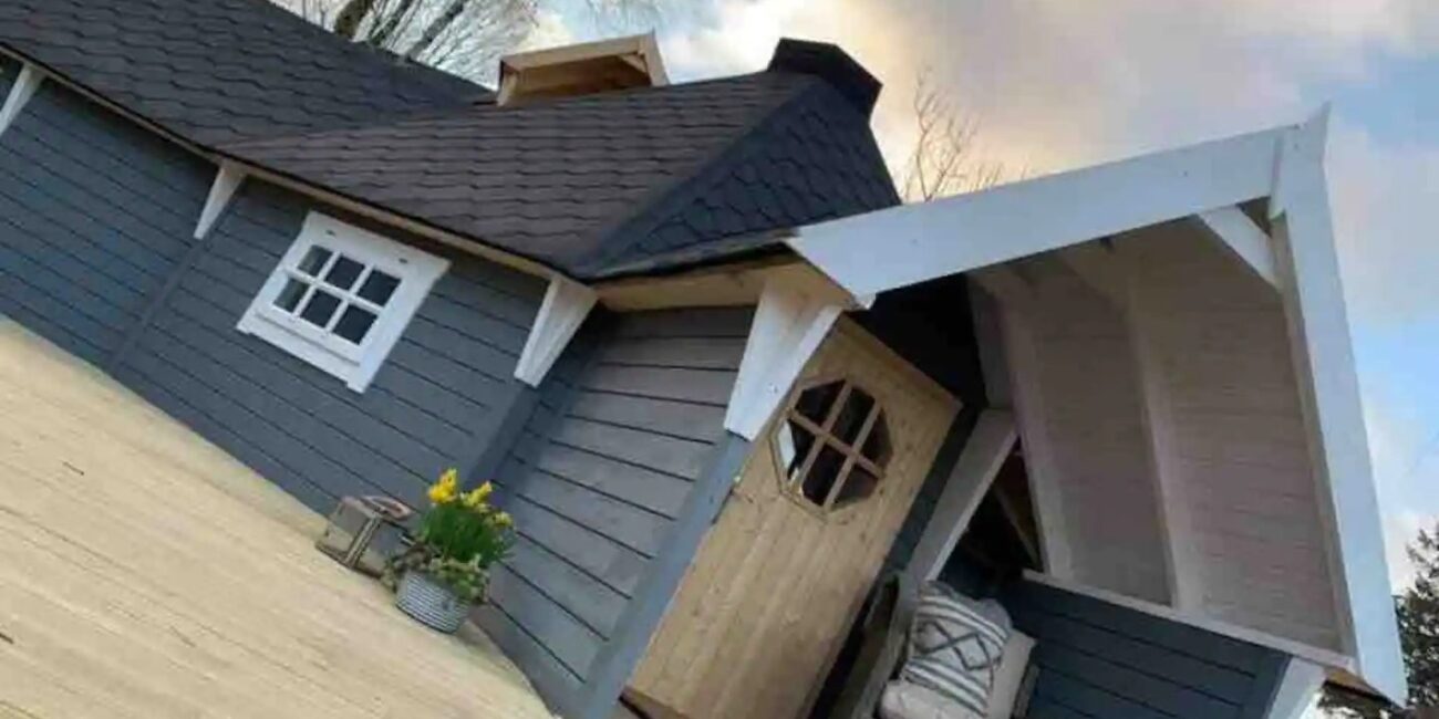 Caban pren llwyd gyda tho crib, drws pren a mynedfa dan do yn Ein Trysor Cudd.