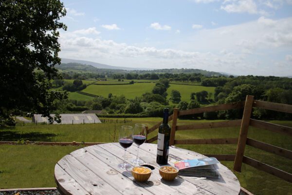 Wine and an open book on a round wooden table beside a fence, overlooking green fields and distant hills at Seren Bach.
