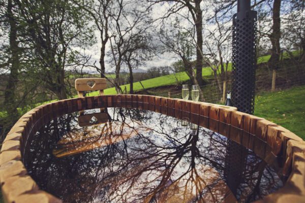 Twb poeth pren crwn yn adlewyrchu coed cyfagos, gyda simnai’r stôf a chaeau gwyrdd y tu ôl.