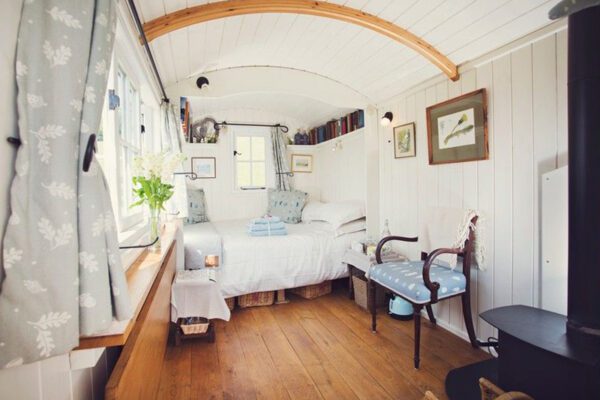 Double bed, patterned armchair and wood-burning stove inside the white-panelled Shepherd’s Hut.