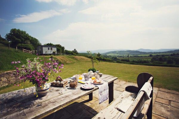 Breakfast laid on an outdoor wooden table with flowers, with the Shepherd’s Hut across the lawn and rolling countryside beyond.