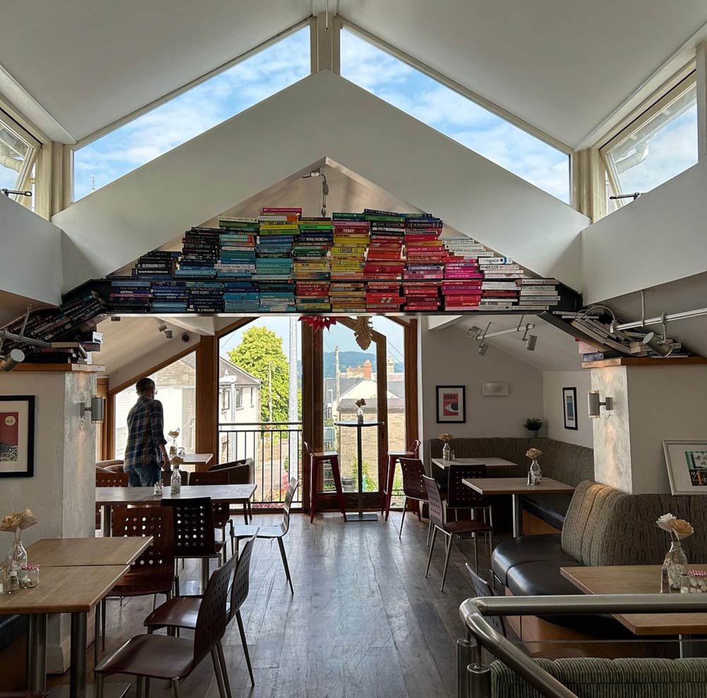 Tables and chairs beneath a pitched roof with a display of colourful books in the Book-ish Café loft.