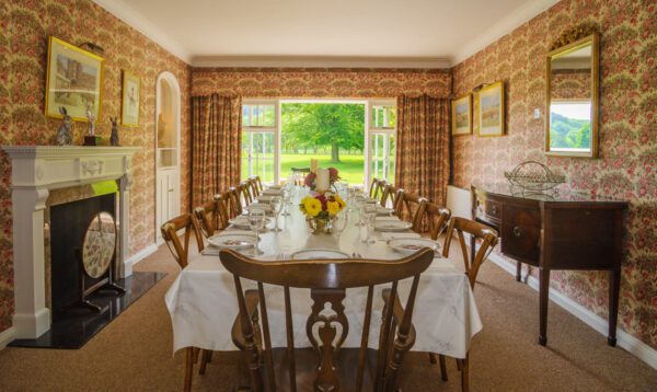Long dining table laid for a meal, with floral wallpaper and a view onto the garden at Glanusk Lodge