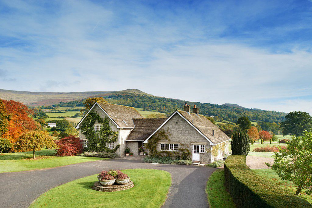 Stone house with climbing plants, a curved driveway and gardens, with wooded hills behind Glanusk Lodge