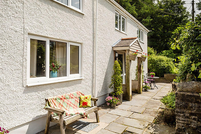 The white-rendered Barn Cottage with a timber porch, paved path, bench and flowering pots.
